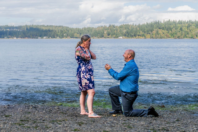 Surprise proposal photography on a Puget Sound beach in Washington as a man kneels at the water's edge