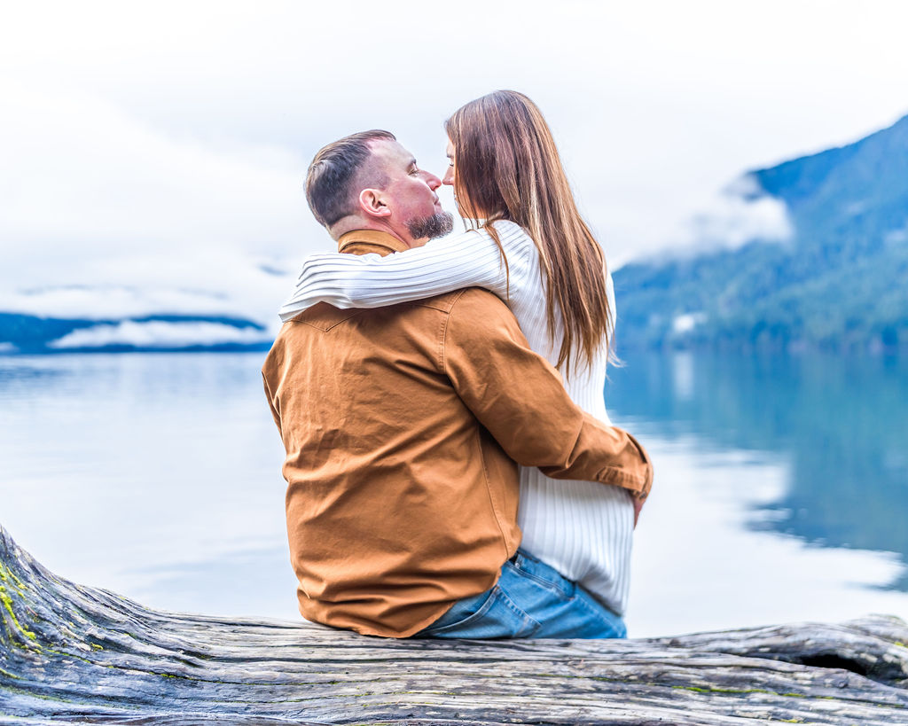 Engaged couple embracing while sitting on a lakeside log during their engagement session at Lake Crescent in Olympic National Park.