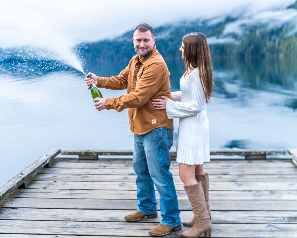 Couple celebrating their engagement with a champagne pop on the dock at Lake Crescent in Olympic National Park.