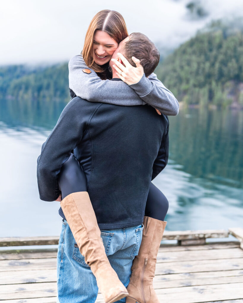 Couple celebrating their engagement with a joyful embrace on a dock at Lake Crescent in Olympic National Park.