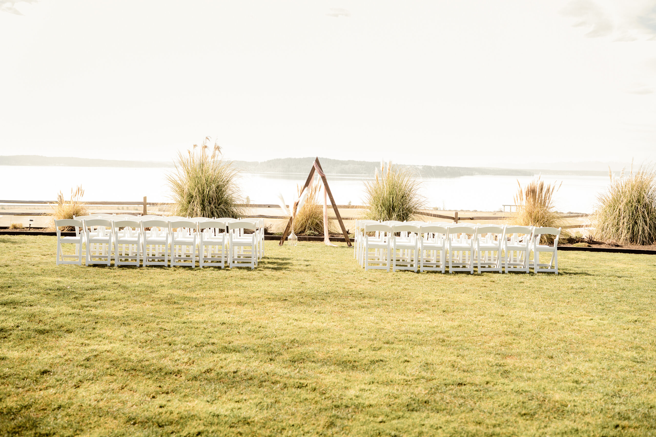 Waterfront wedding ceremony space with a triangular arch and rows of white chairs on a lawn overlooking the water