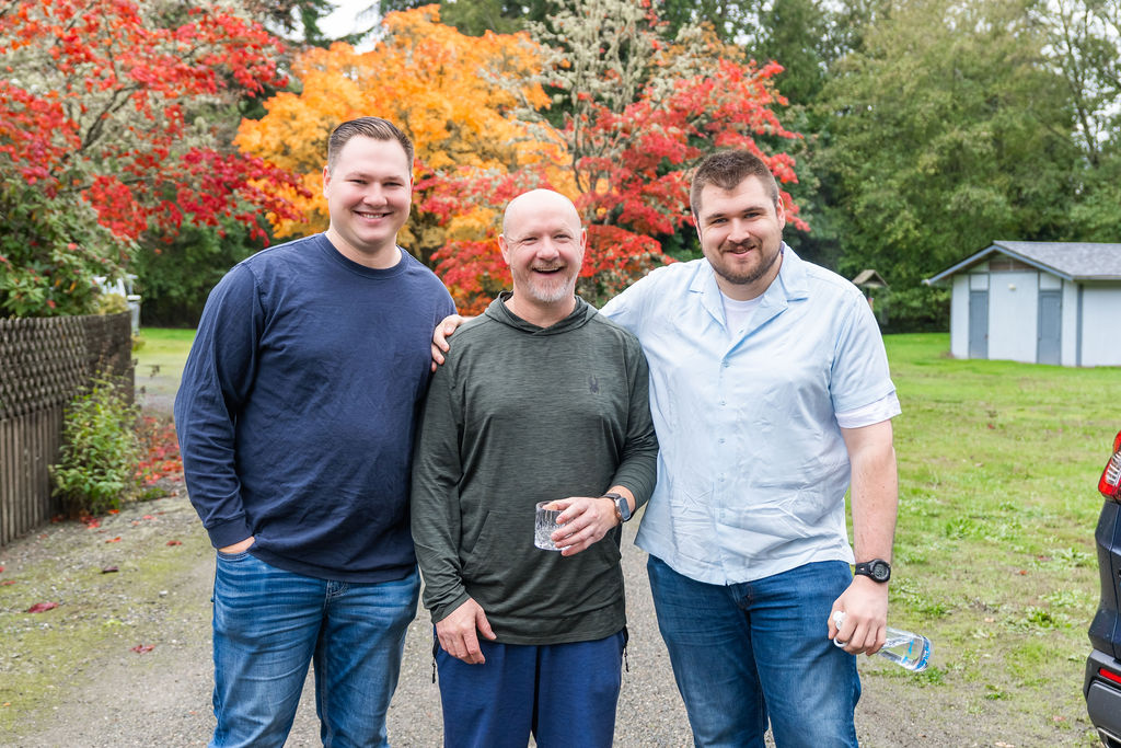 Three men smiling together outdoors during a Vashon wedding with colorful fall trees in the background