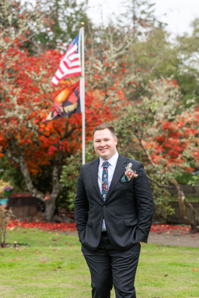 Groomsman in a black suit standing in front of colorful autumn trees and American flag at a Vashon Island wedding in Washington