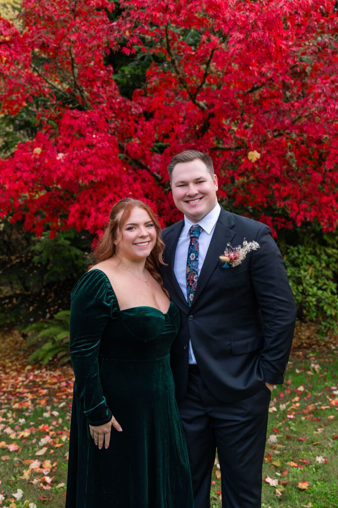 Smiling wedding couple posing together in front of bright red autumn trees during a fall wedding on Vashon Island, Washington