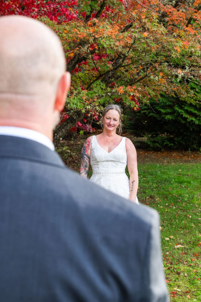 Bride smiling during a first look moment as the groom turns to see her beneath colorful fall trees at a Vashon Island wedding