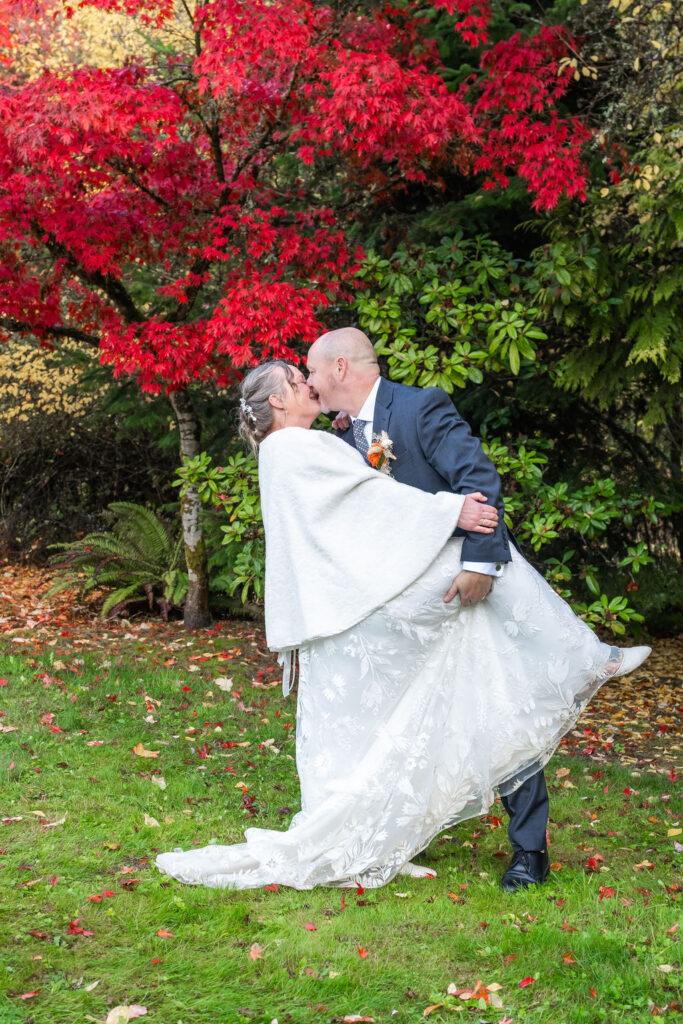Groom dipping bride for a kiss beneath vibrant red autumn trees during a fall wedding portrait on Vashon Island, Washington