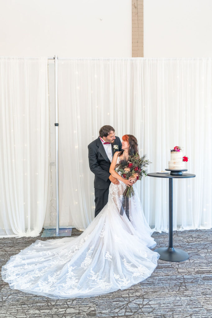 Newlywed couple posing near wedding cake inside Vickery Gardens reception hall
