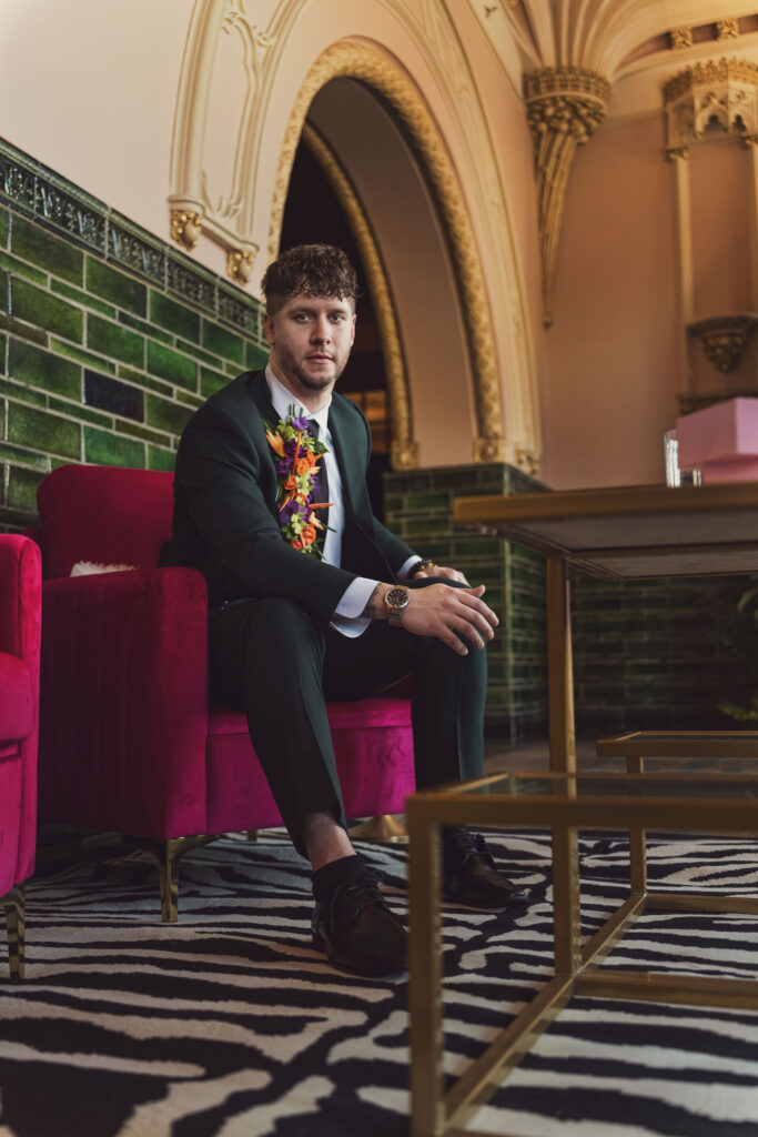 Groom in a green suit with a tropical boutonniere seated on a pink velvet chair in an ornate venue