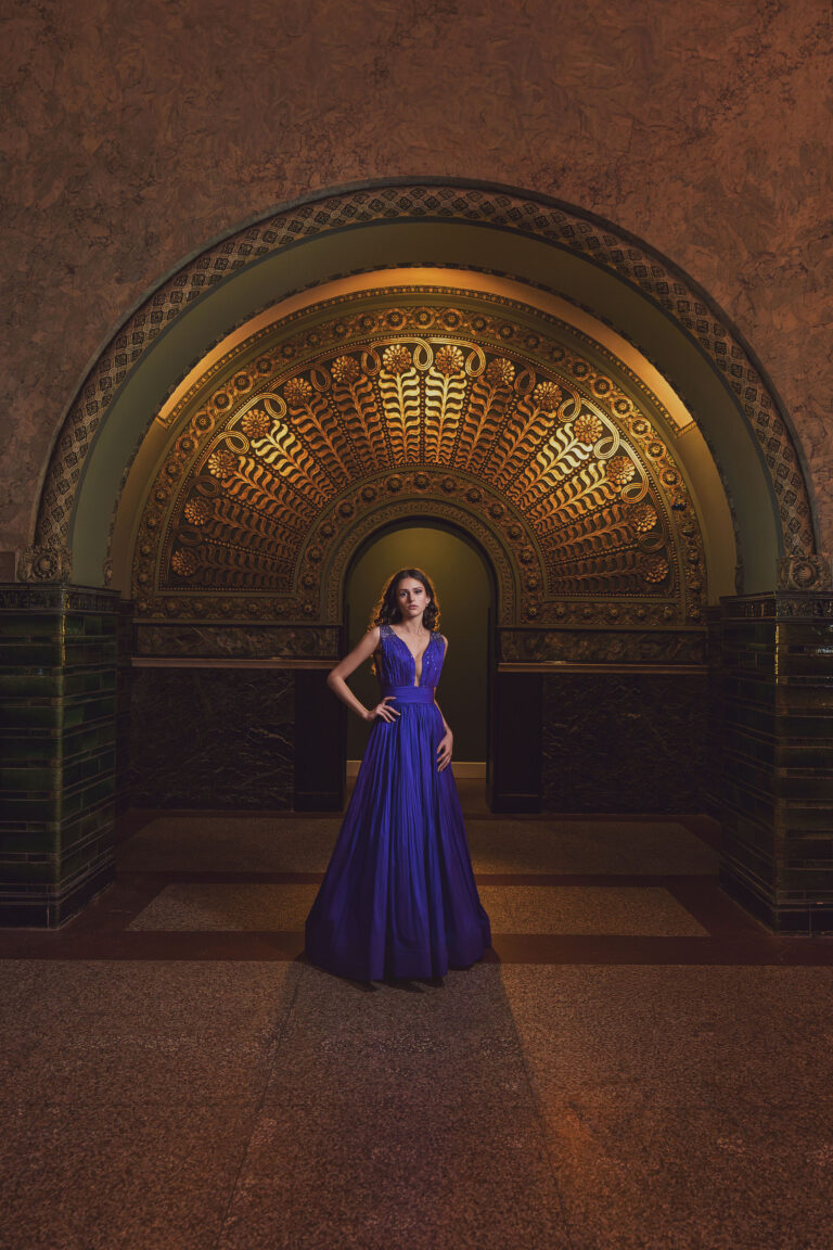 Senior portrait of a young woman in a royal blue gown standing beneath an ornate gold arched ceiling