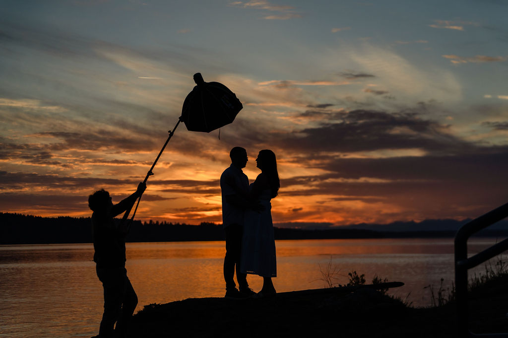 Sunset silhouette of an engaged couple by the water as a photography assistant holds a lighting umbrella, by a Puget Sound wedding photographer