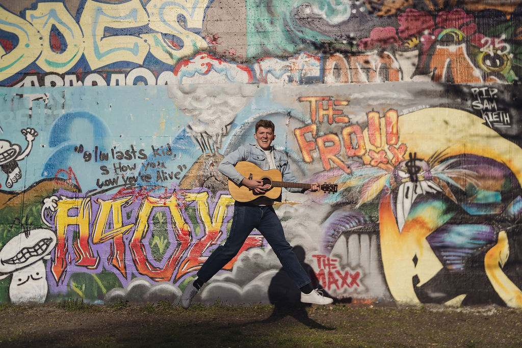 Senior jumping with guitar in front of graffiti wall.