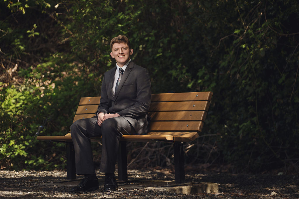 Kitsap County senior photographer portrait of a high school senior in a gray suit seated on a wooden park bench surrounded by greenery
