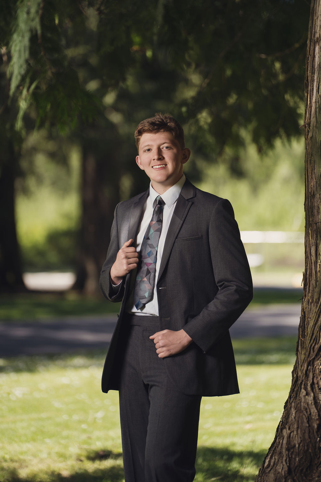 High school senior in a gray suit standing beside a tree in a sunny park