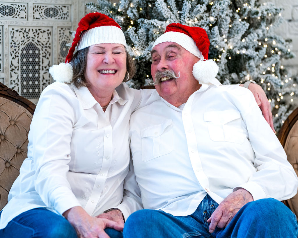 Older couple in Santa hats laughing with eyes closed on a vintage settee in front of a flocked Christmas tree during a Christmas studio session in Bremerton