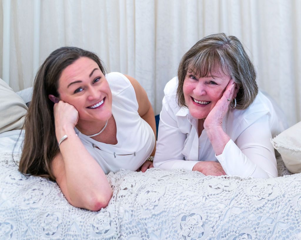 Mother and her adult daughter resting their chins on their hands and laughing on a white bed during a holiday studio session in Bremerton
