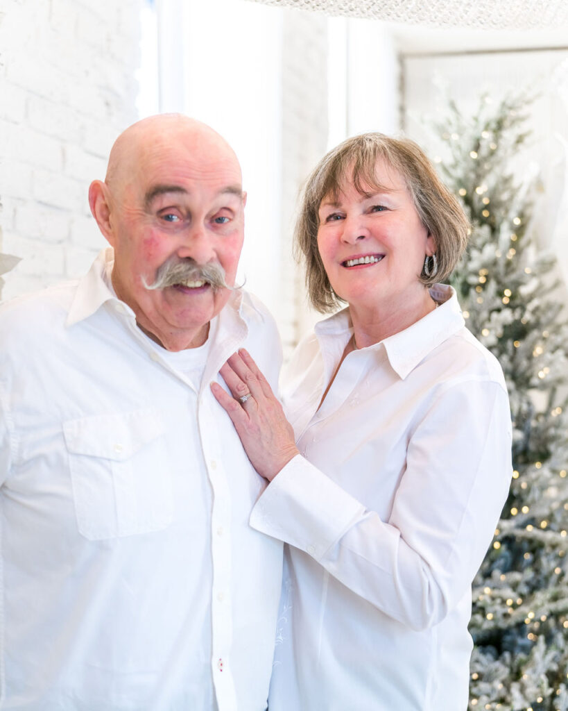 Older couple in white shirts standing together in front of flocked Christmas trees during a holiday studio session in Bremerton