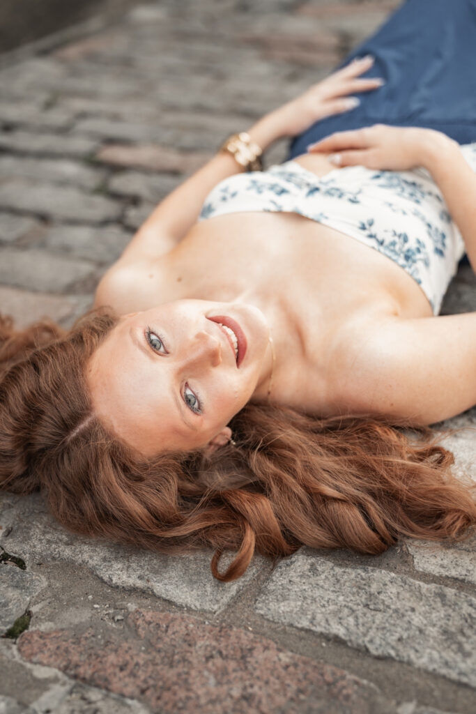 Creative senior portrait of a red haired young woman lying on cobblestones with her hair fanned out