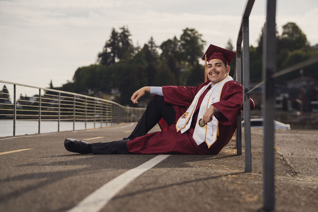 Kitsap County senior photographer photo of a graduate in maroon cap and gown sitting on a waterfront walkway