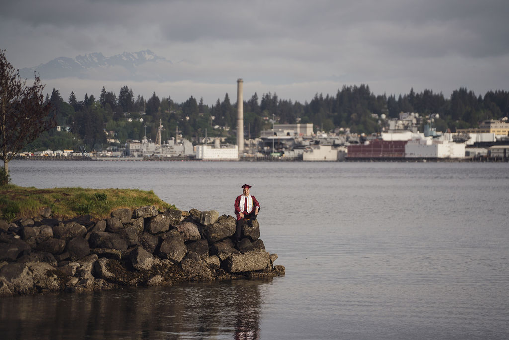 Graduating senior in cap and gown standing on a rocky shoreline with the Puget Sound waterfront and Olympic Mountains behind