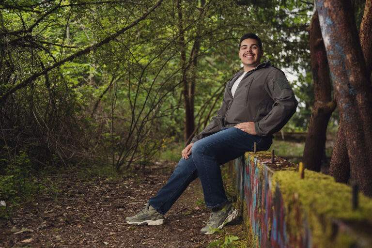 High school senior seated on a mossy barrier in lush green woods during a senior session