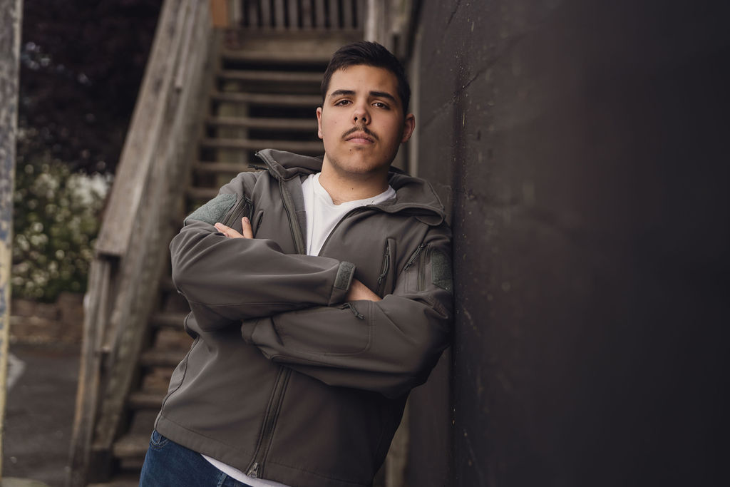 High school senior in a gray jacket leaning against a wall with arms crossed near a wooden staircase