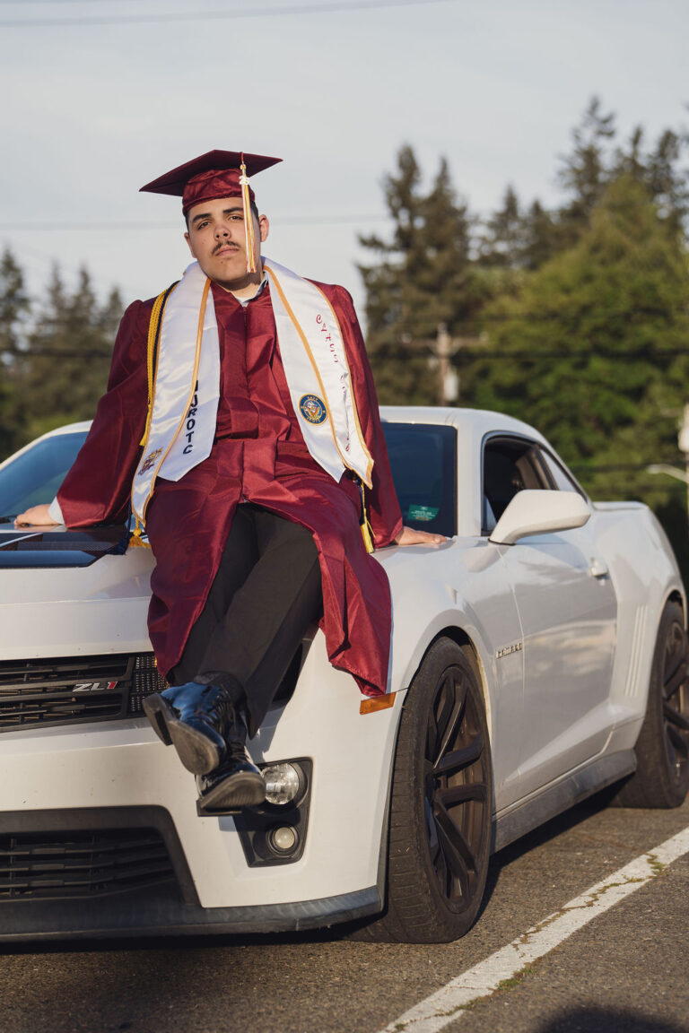 Graduating senior in a maroon cap and gown sitting on the hood of a white Chevrolet Camaro