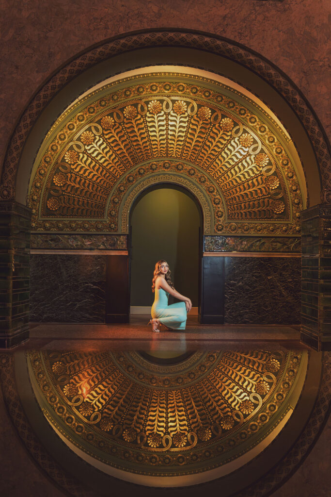 Elegant senior portrait of a young woman in an aqua jumpsuit crouched beneath an ornate gold mosaic arch with a mirrored reflection