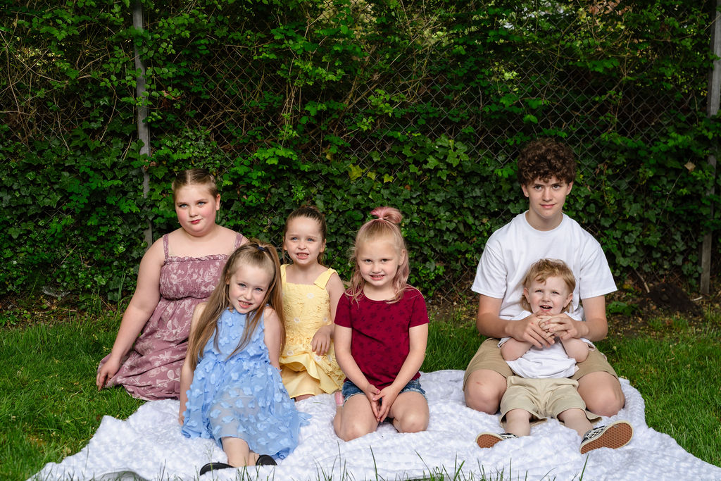Six cousins sitting together on a white blanket in front of leafy greenery at a Kitsap County park