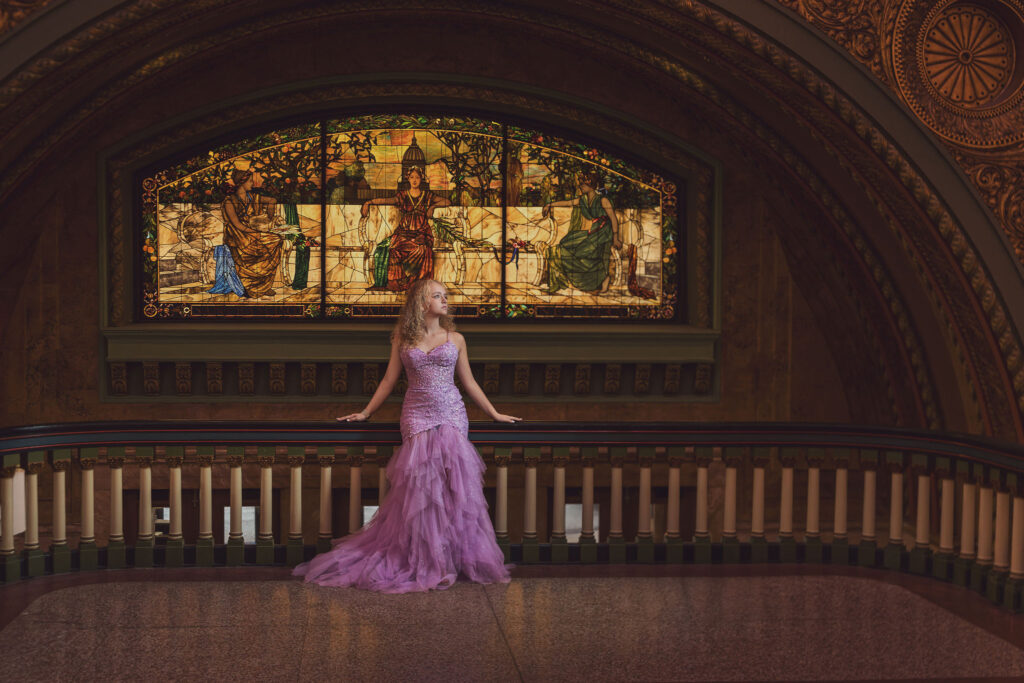 Formal senior portrait of a young woman in a lavender gown before a grand stained glass window in a historic hall