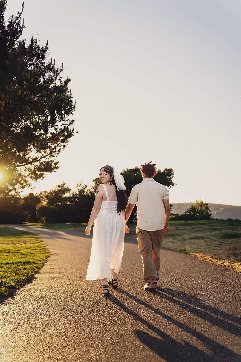 Engaged couple walking hand in hand down a path as the bride to be glances back with the sunset behind them at Chambers Bay near Tacoma