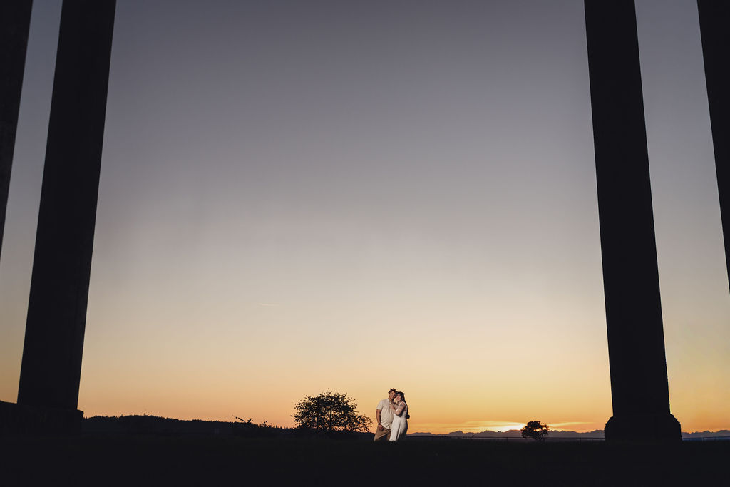 Engaged couple embracing in silhouette between the towering concrete columns at Chambers Bay during a vivid sunset near Tacoma