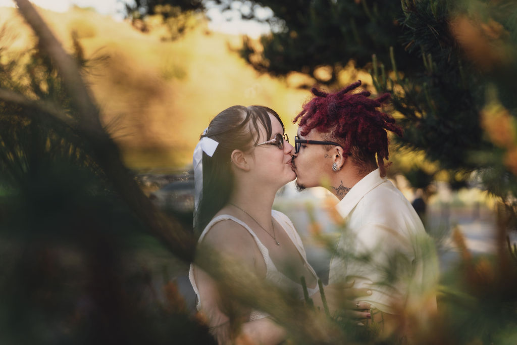 Engaged couple kissing at golden hour framed through pine branches at Chambers Bay near Tacoma