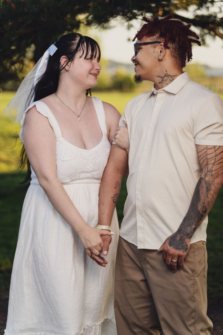 Engaged couple smiling at each other while holding hands in a sunlit meadow at Chambers Bay near Tacoma