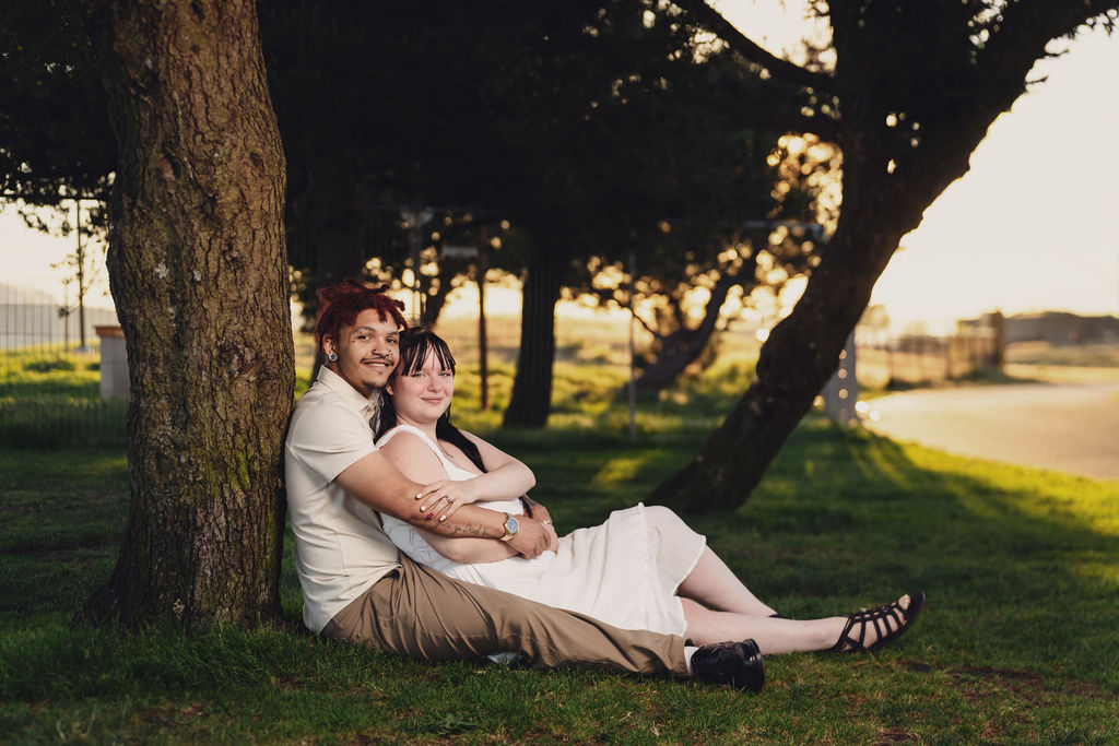 Engaged couple relaxing against a tree in the grass at golden hour at Chambers Bay near Tacoma
