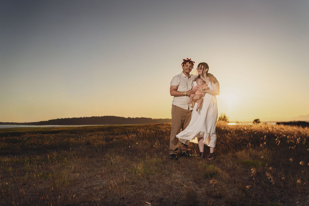 Engaged couple holding their infant daughter in a golden meadow at sunset at Chambers Bay near Tacoma