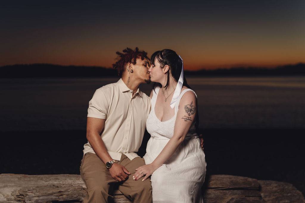 Engaged couple sharing a kiss while seated on driftwood at twilight at Chambers Bay near Tacoma