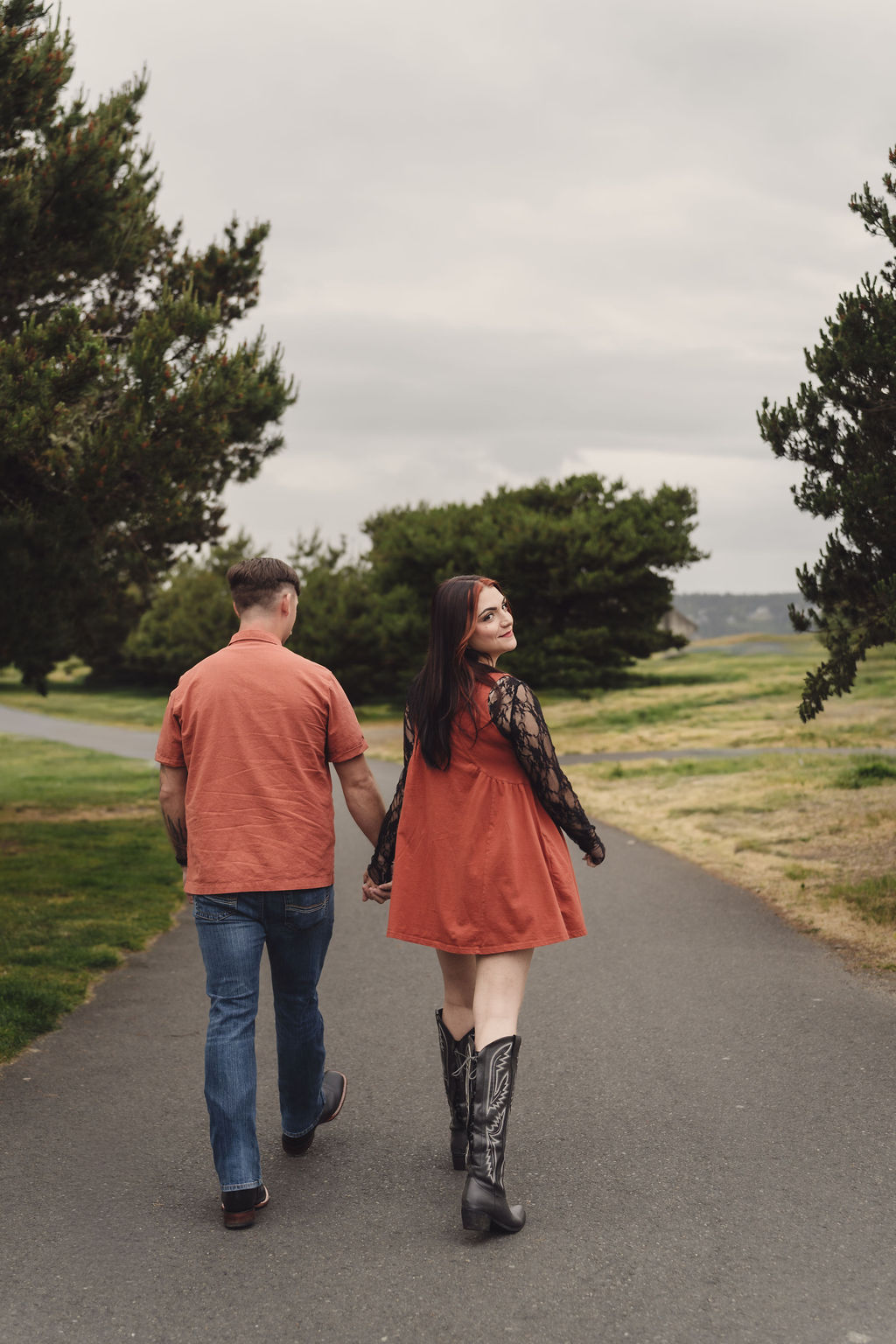 Engaged couple walking hand in hand down a path at Chambers Bay as the bride to be glances back over her shoulder near Tacoma
