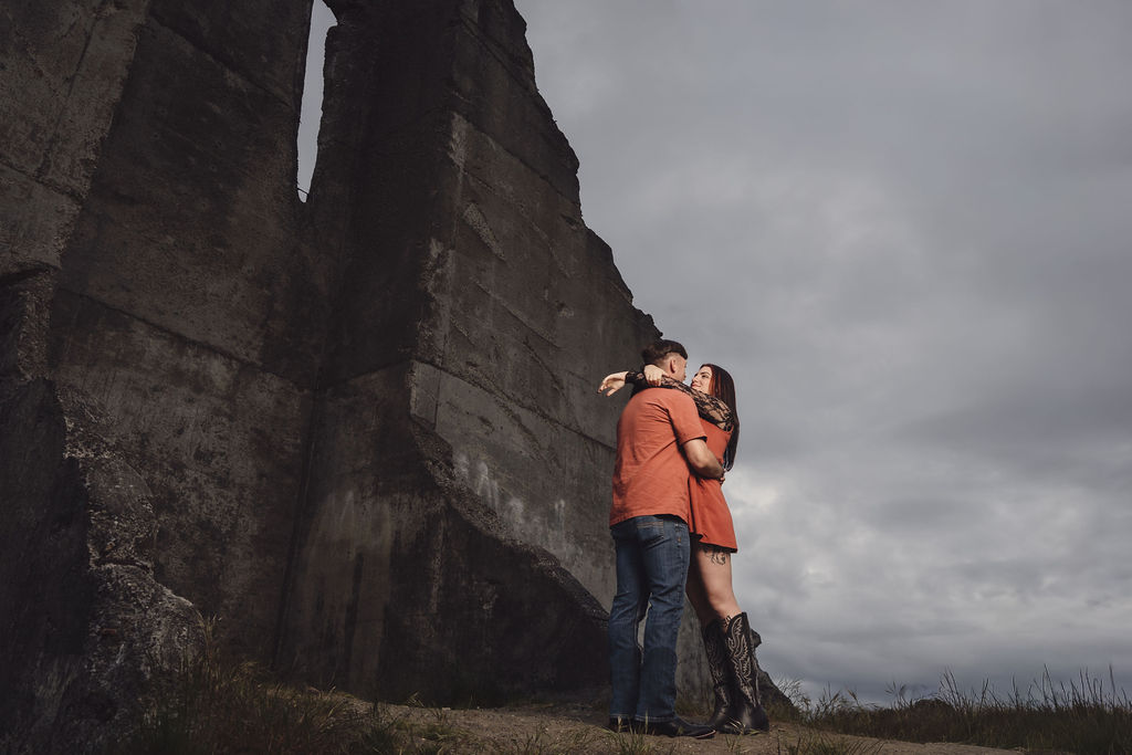 Engaged couple embracing at the base of the towering concrete ruins at Chambers Bay near Tacoma under a moody sky