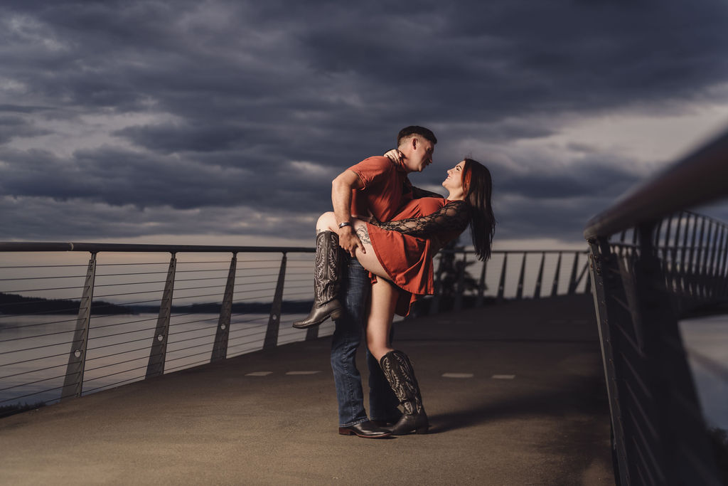 Partner dipping his fiancee on the boardwalk pier over the water at Chambers Bay under dramatic clouds near Tacoma