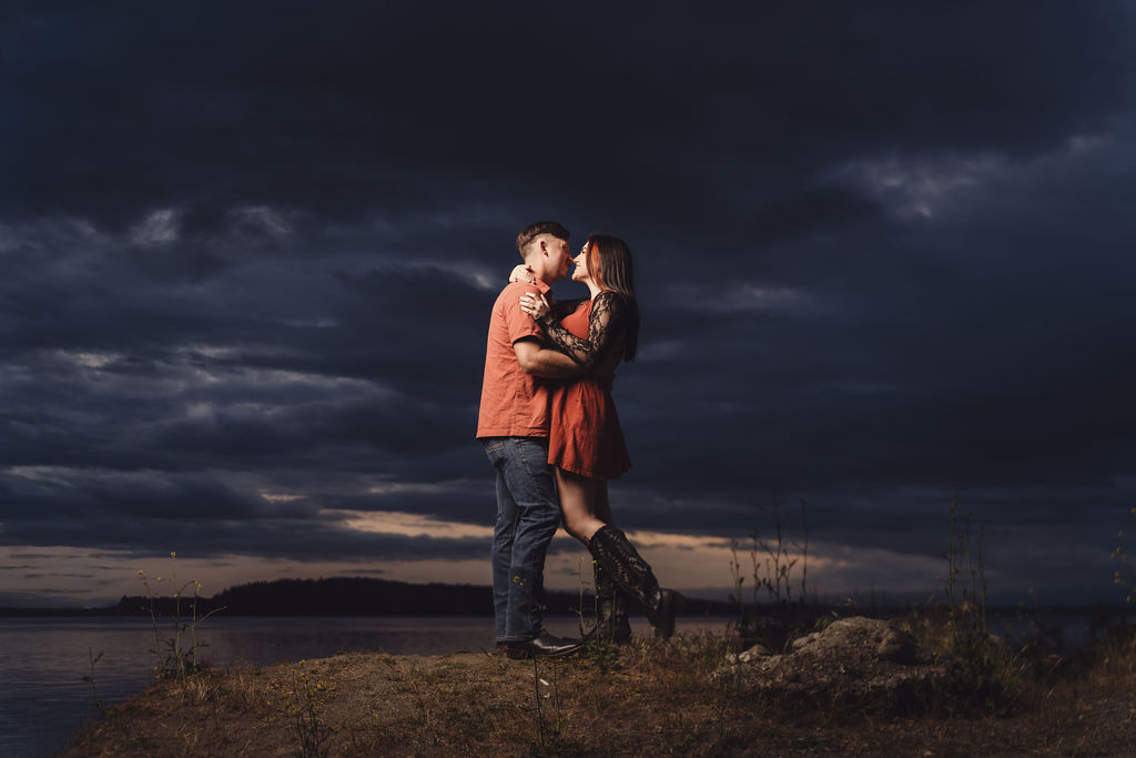 Engaged couple embracing on a bluff above the water under dramatic dark clouds at Chambers Bay near Tacoma