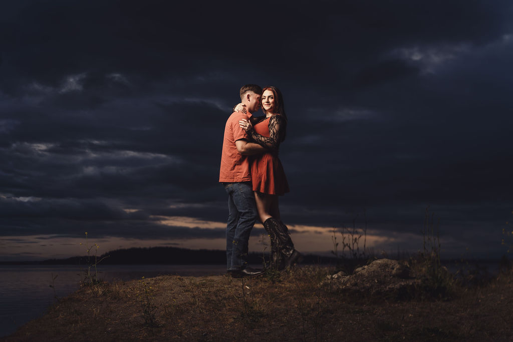 Engaged couple embracing on a bluff above the water at Chambers Bay under a dark dramatic sky at dusk near Tacoma