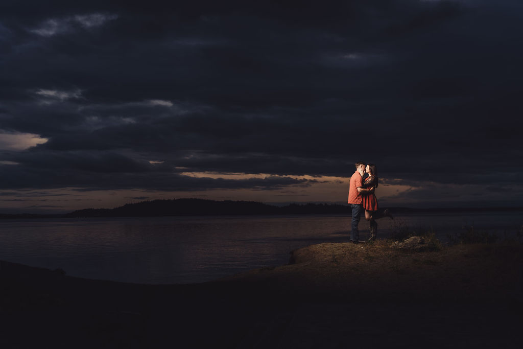 Engaged couple standing together on the shoreline at Chambers Bay with a wide moody sky over Puget Sound near Tacoma at dusk
