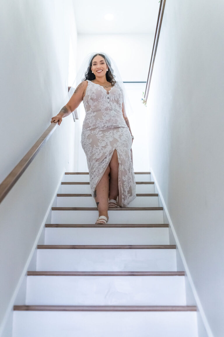 Smiling bride in a lace wedding gown and veil walking down a bright staircase before her ceremony