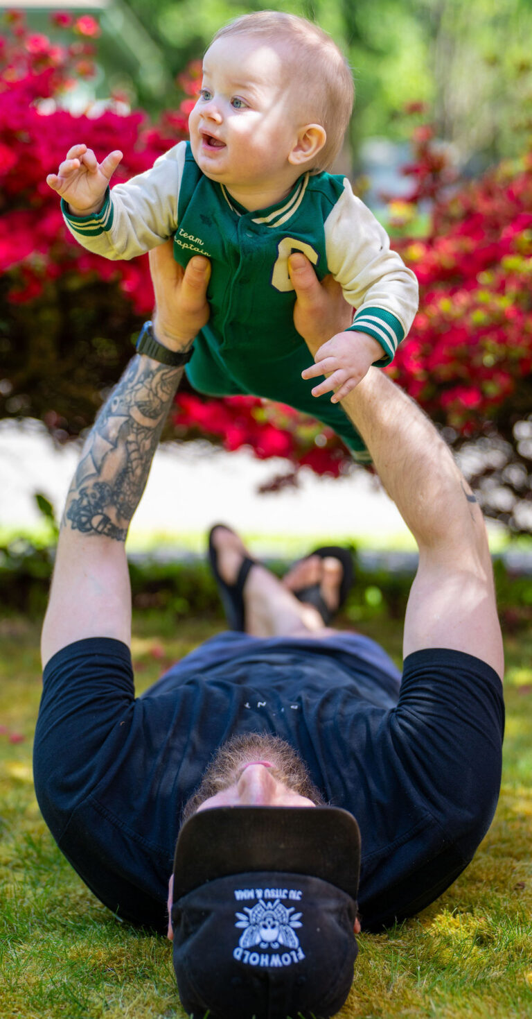 Father lying in the grass lifting his smiling baby overhead with red spring blooms behind them in Kitsap County