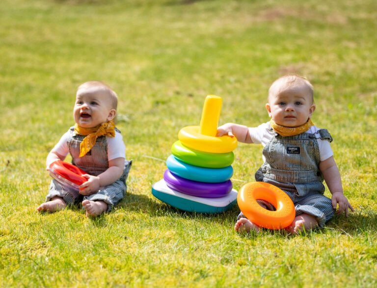 Twin babies in striped overalls playing with a stacking ring toy in the grass during an outdoor family session in Kitsap County