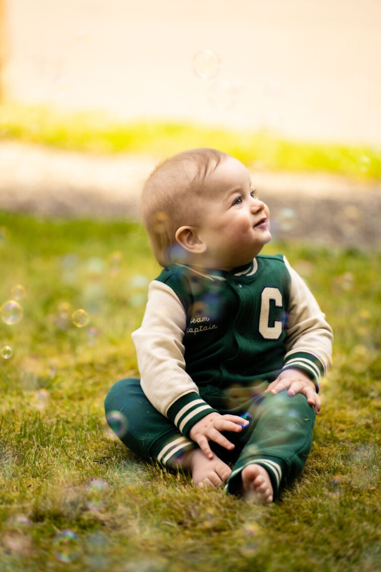 Smiling baby in a green varsity romper sitting in the grass with bubbles floating around in Kitsap County