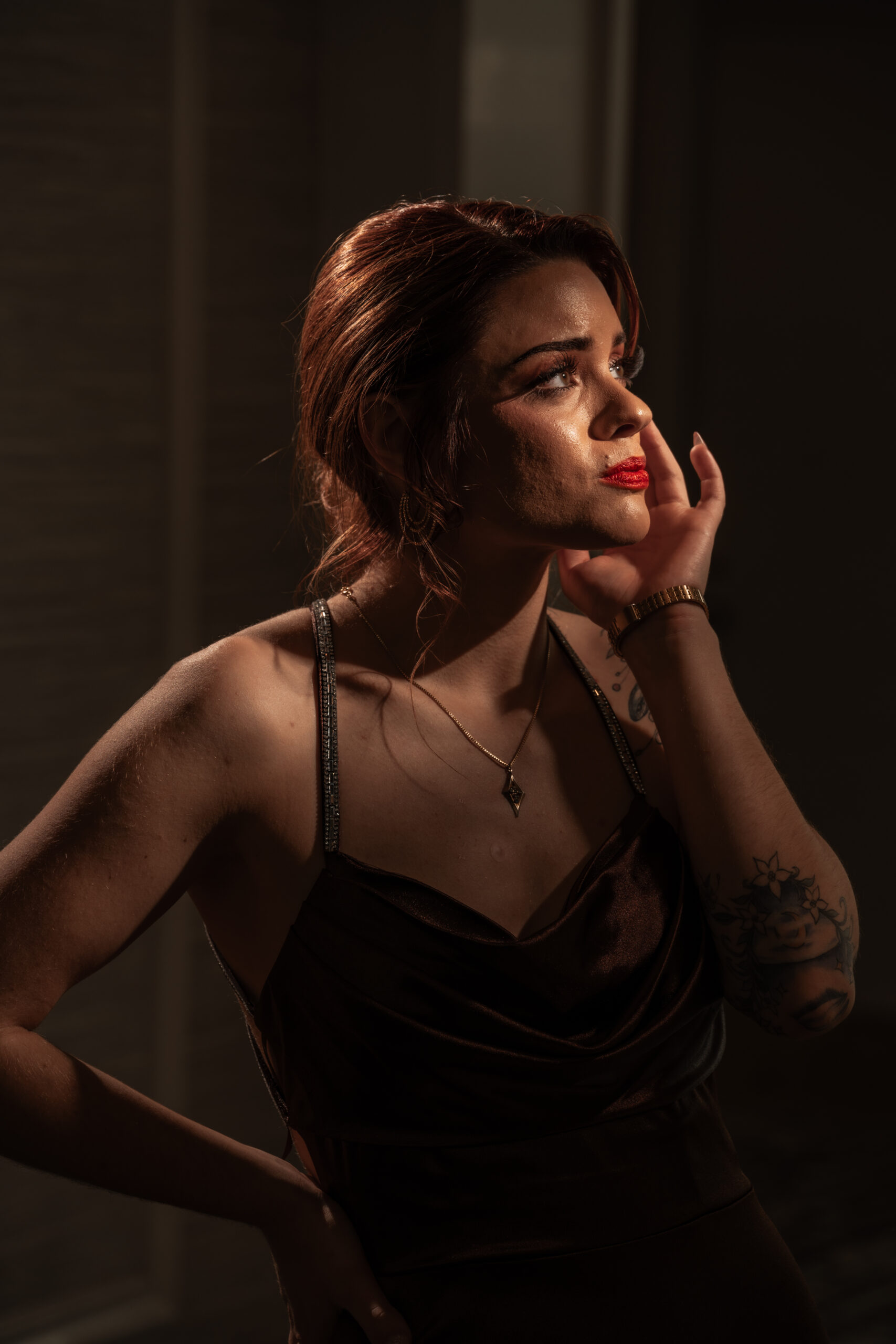 High fashion senior portrait of a young woman in a dark satin slip dress with bold red lipstick in dramatic light