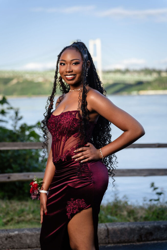 Smiling South Kitsap prom senior in a burgundy gown with a leg slit posing at the Tacoma Narrows Bridge overlook