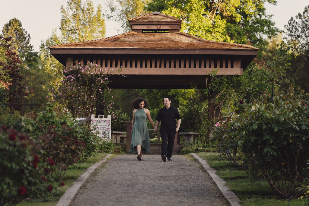 RJ and Maya walking together through an open grassy area at Point Defiance Park during their engagement session