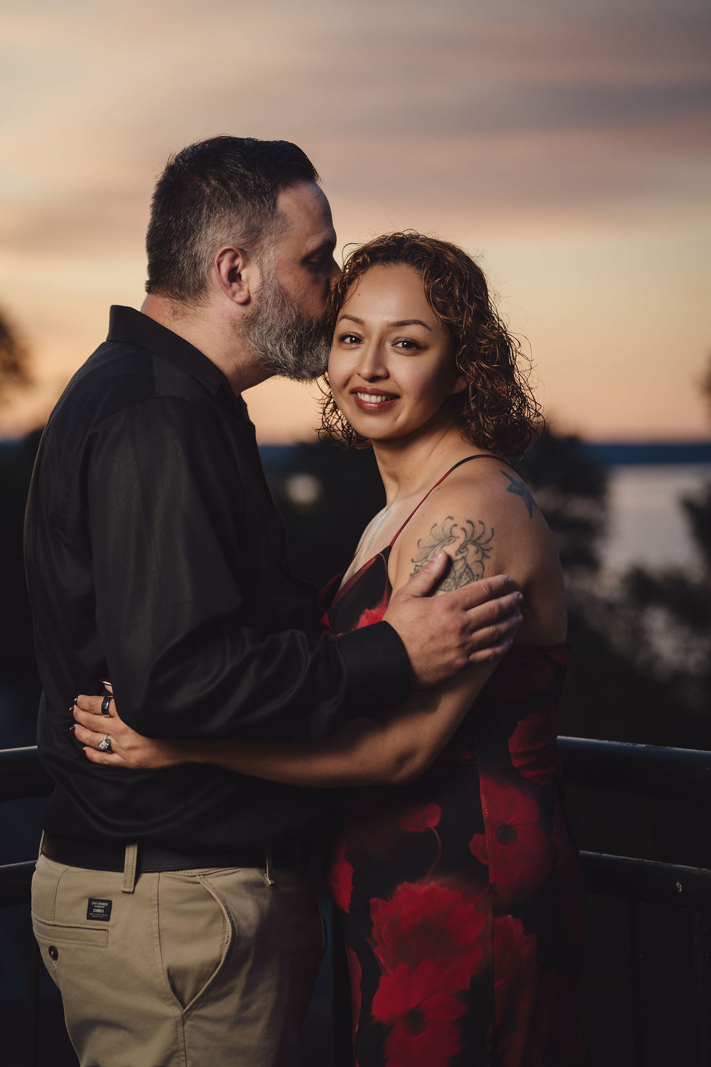 Partner kissing his fiancee on the head while she smiles with Commencement Bay behind them in Tacoma at sunset